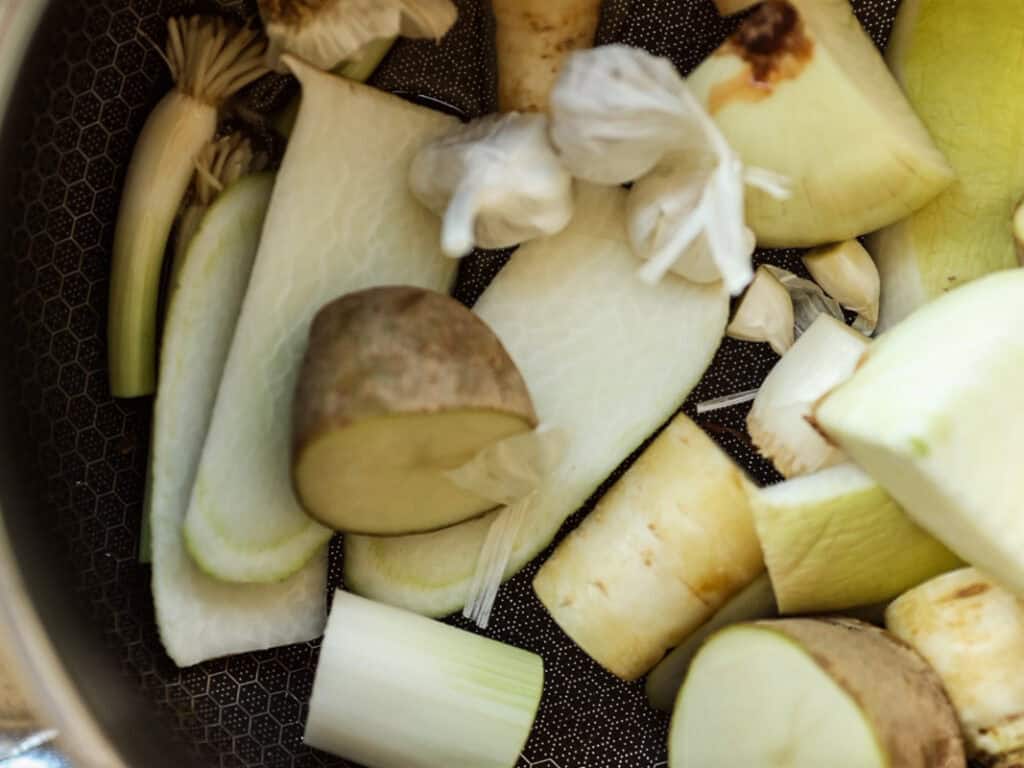 A close-up of various chopped vegetables in a pan, including potatoes, onion, garlic, leek. The ingredients are in preparation for cooking, with visible skin and varied textures in a black pan.